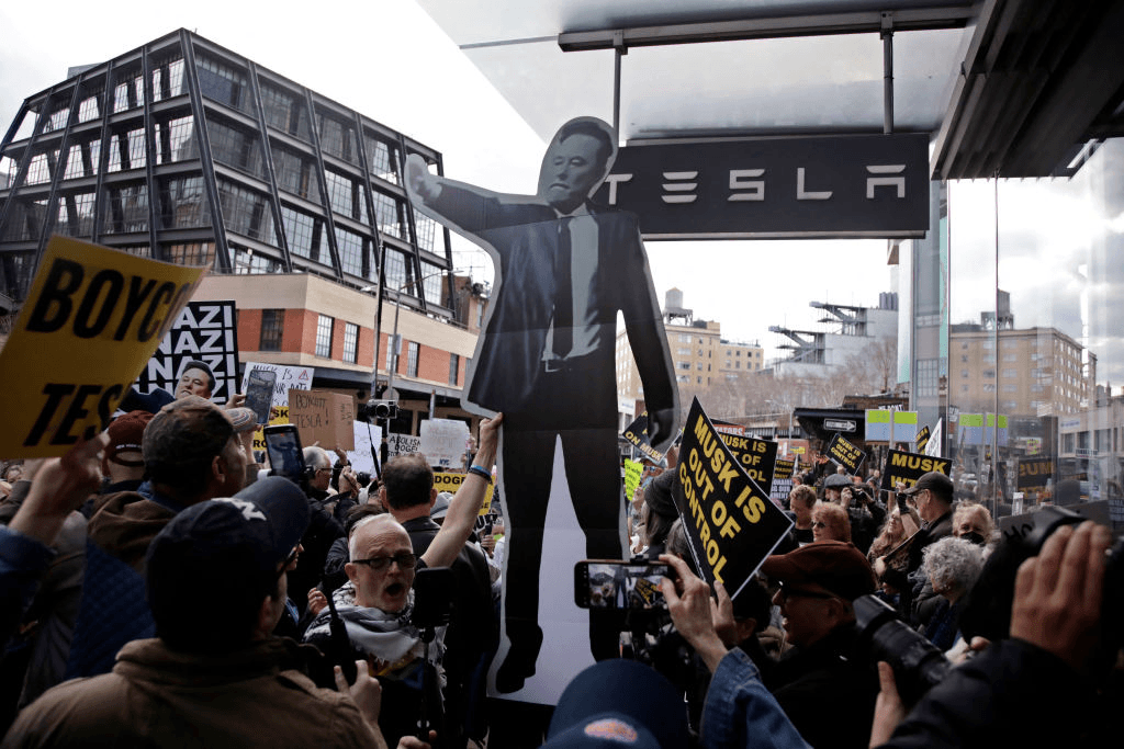 crowd outside tesla shop holding placards saying "nazi" and a cut-out of elon musk doing the nazi salute.