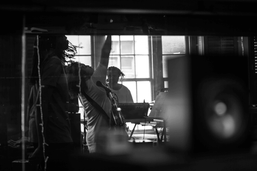 Black and white photo of musicians in a studio. One person is energetically playing guitar with their arm raised, while another stands by a keyboard, visible through a window. A speaker is in the foreground. The scene conveys dynamic musical activity.
