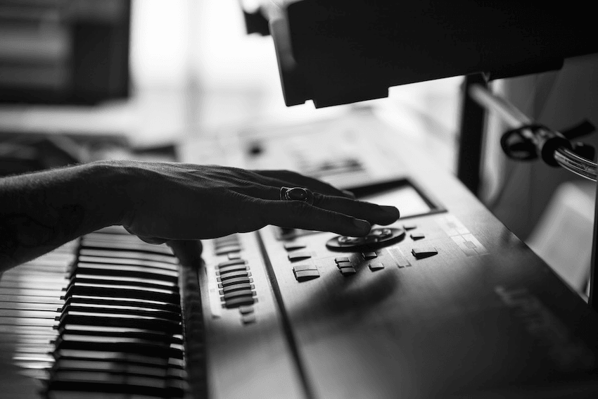 A black and white image of a hand adjusting controls on an electronic keyboard or synthesizer. The fingers appear to be fine-tuning a knob or button, with the keyboard keys visible in the foreground. The scene is softly lit.