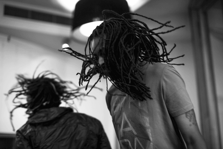 Two people with dreadlocks dance energetically in a dimly lit room. One wears a T-shirt, the other a jacket. Their hair swings dramatically with their movements. The background is softly blurred, focusing on their dynamic motion. Black and white image.