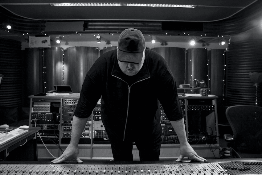 A person in a cap leans over a mixing console in a dimly lit recording studio. The space is filled with audio equipment and illuminated by soft lights. The person is focused on the controls and wears a dark jacket.