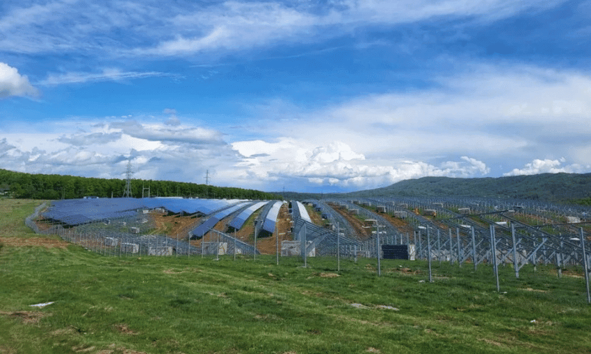 A vast green field with rows of solar panels under a blue sky with scattered clouds. Mountains and trees are visible in the background.