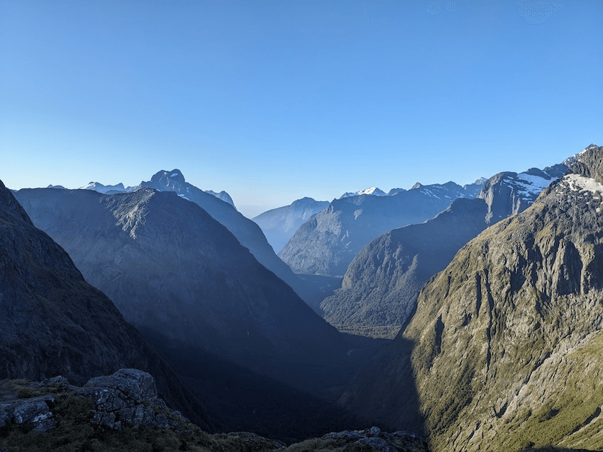 shadows fall between blue, steep mountains in the evening sunlight, with a twining flat glimpse of a fjord beyond