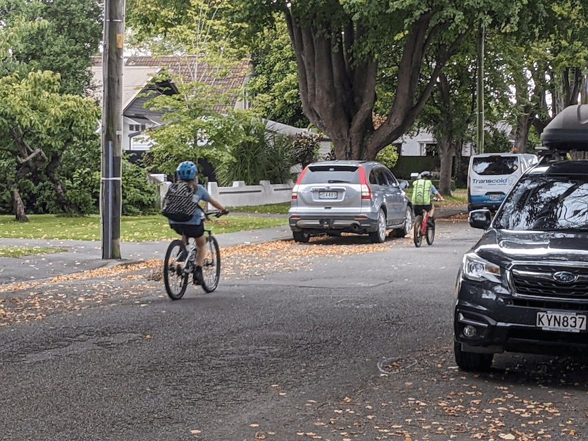 a suburban street with parked cars and big trees and two kids cycling