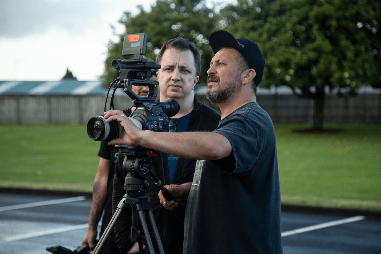 Two men are outside in a parking lot, focused on using a professional video camera mounted on a tripod. One is adjusting the camera while the other looks on. A tree and some buildings are visible in the background.