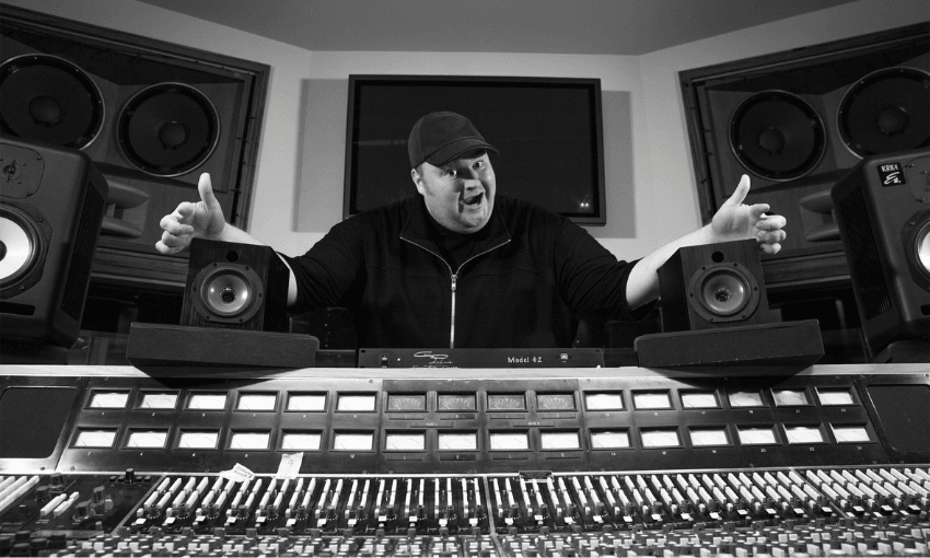 A person wearing a cap and jacket stands behind a large audio mixing console in a recording studio, arms spread wide. Speakers and a screen are visible in the background. The image is in black and white.