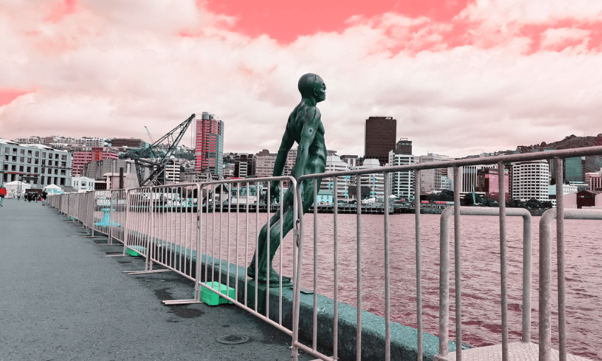 A bronze sculpture of a man appears to be diving into the water, positioned on a waterfront with railings. Urban buildings and construction cranes are visible across the water under a cloud-filled sky with a pinkish hue.