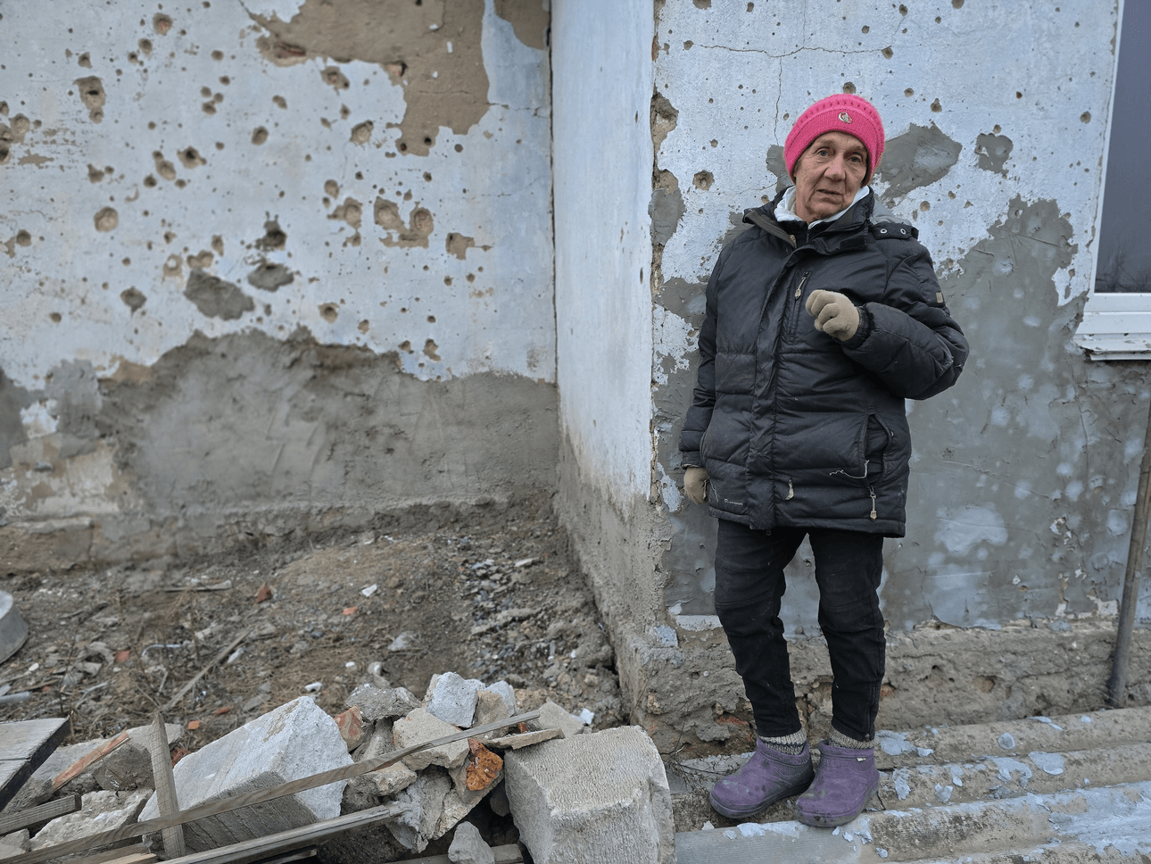 A woman in a pink beanie and puffer jacket stands in front of a home riddled with bullet holes
