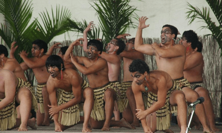 A group of performers in traditional attire, with faces painted in moko kanohi, perform a haka. They have bare chests and wear piupiu skirts made of harakeke flax. Palm plants are in the background. The scene is dynamic, showcasing strong gestures and facial expressions.