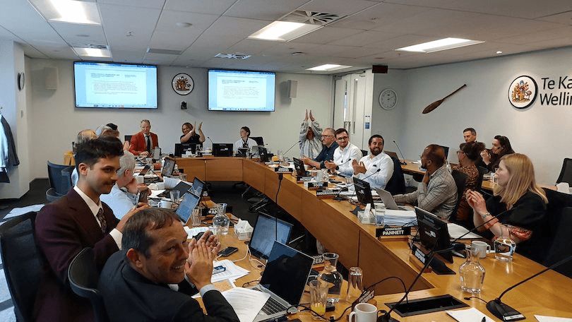 A group of people in a conference room sit around a large table, engaged in discussion. Laptops and documents are in front of them. Two screens display content on the wall. The atmosphere appears collaborative and focused.