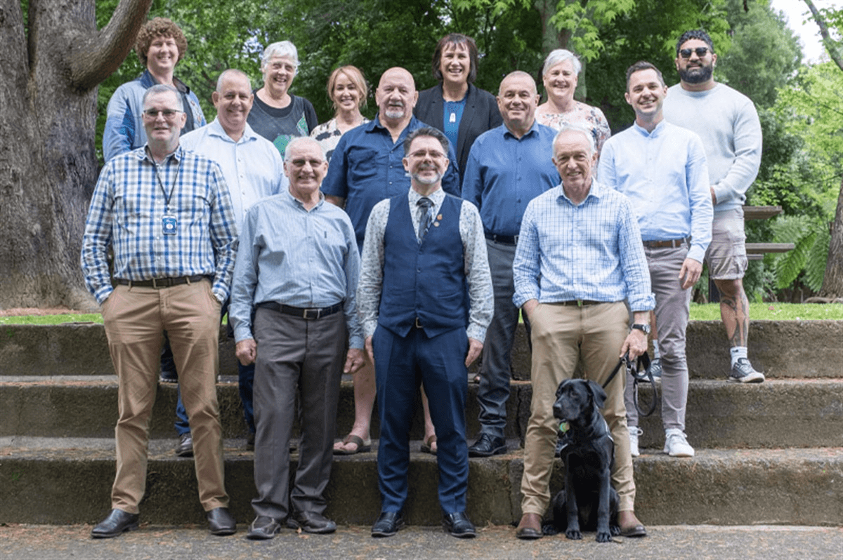 A group of 14 people pose on outdoor steps, smiling at the camera. One person has a black dog. They are in a park with trees in the background. The group includes a mix of men and women in casual and business casual clothing.