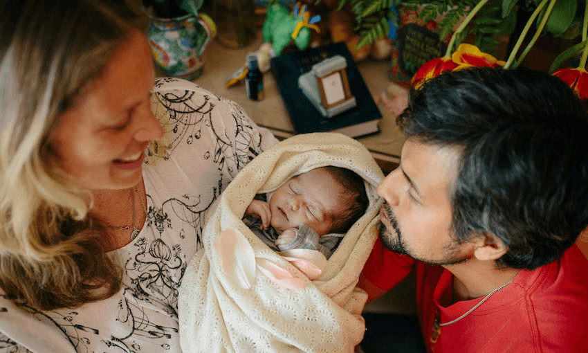 A smiling woman and man admire a newborn wrapped in a cream-colored blanket. The baby sleeps peacefully. Colourful flowers and a few items are blurred in the background.