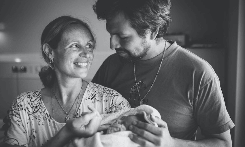 A couple lovingly looks at each other while holding a newborn wrapped in a blanket. Both are smiling, standing closely, with soft lighting creating an intimate atmosphere. The image is in black and white.