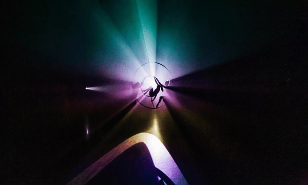 A silhouetted performer spins inside a circular apparatus in a sequence from Belle - A Performance on Air at Te Ahurei Toi o Tāmaki Auckland Arts Festival, surrounded by vibrant beams of colourful light in a dark space.