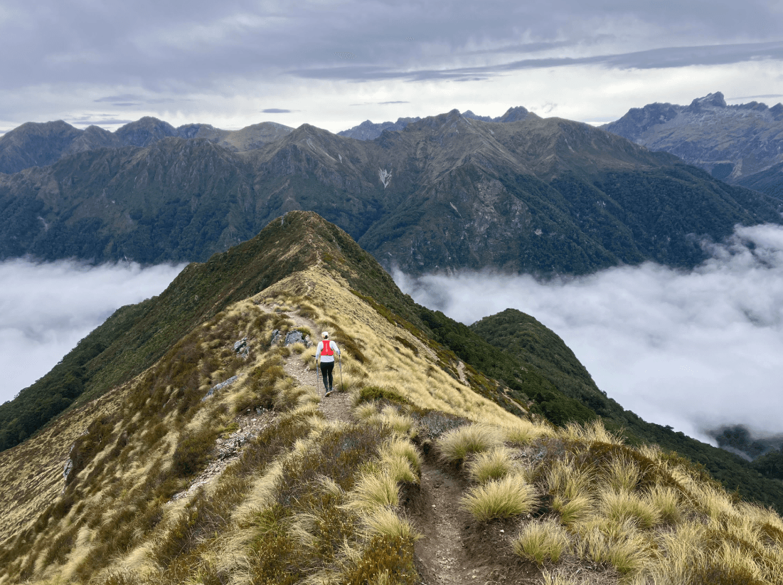 a small figure hikes mountain ridge with clouds below