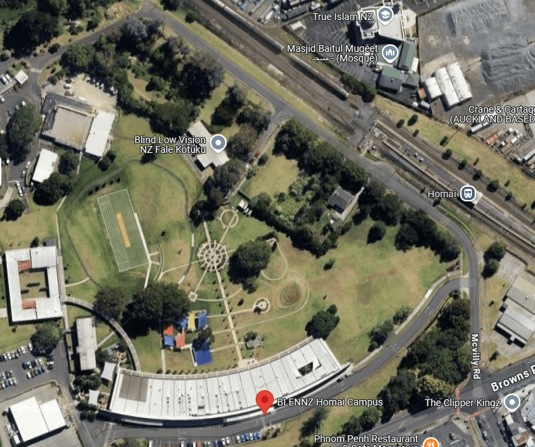 Aerial view of a campus with a circular garden and green space in the center, surrounded by roads and buildings. To the left are sports fields and a track. A railway line runs across the top, and nearby buildings are labeled with various names.