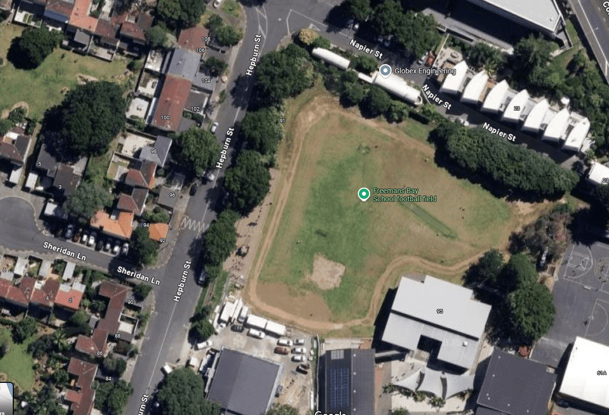 Aerial view of a rectangular sports field surrounded by houses and trees. Roads and buildings are visible around the field, with trees lining its perimeter. The label "Greenland Bay School Football Field" is marked on the field.