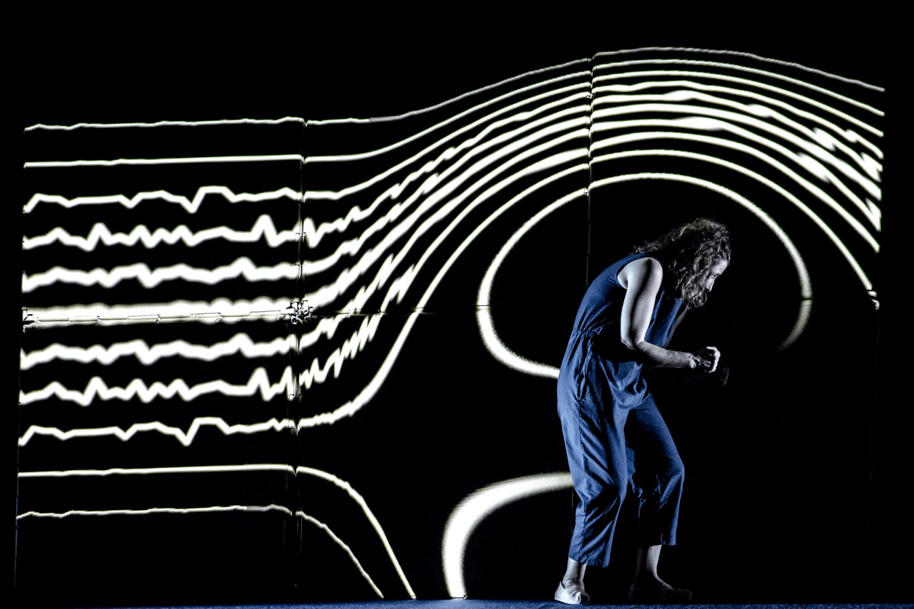 a woman on a dark stage in a blue jumpsuit. Behind her a pattern of white lines are projected, seemingly curing around her.