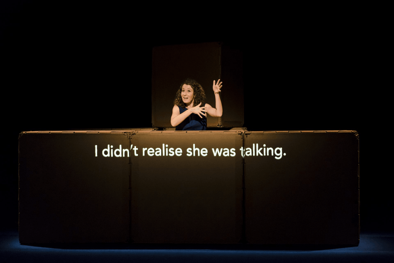 woman on a dark stage talking with sign language. She is leaning on boxes upon which there is a projection reading "I didn't realise she was talking". 