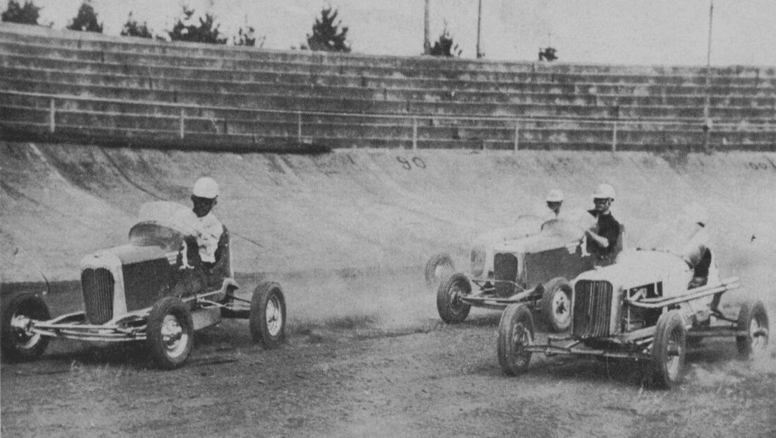 Three vintage race cars, each with a driver wearing a helmet, speed around a dirt track on a banked curve. The grandstand in the background is empty, and trees are visible behind the seating area. Dust trails behind the cars, adding a dynamic feel.