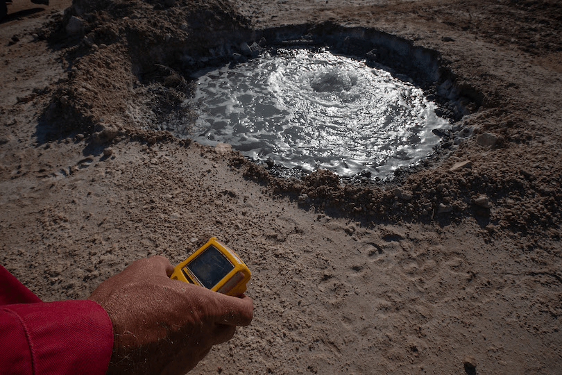 A person holds a yellow device near a small bubbling geothermal pool on rocky terrain. The pool has a circular motion, indicating hot water activity. The landscape looks arid, with dry soil around the pool.