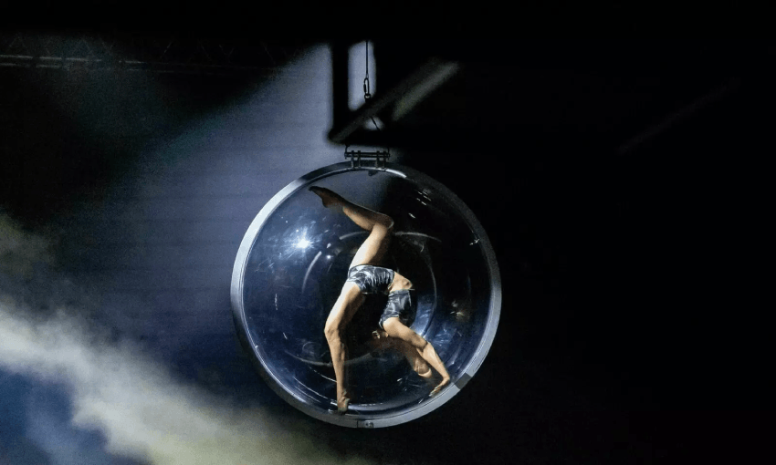 A performer is curled up inside a large, transparent sphere suspended in the air. They are wearing a silver and black outfit, and the scene is dimly lit with mist or smoke surrounding the sphere, set against a dark background.