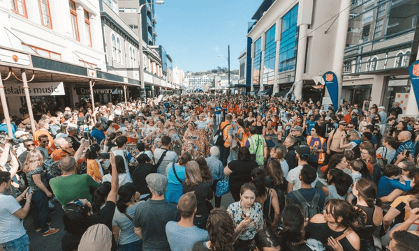 A large crowd gathers on a city street, celebrating with music and dancing. Bright buildings line the street, and the sky is clear. People are dressed casually, some holding cameras and phones, capturing the lively atmosphere.