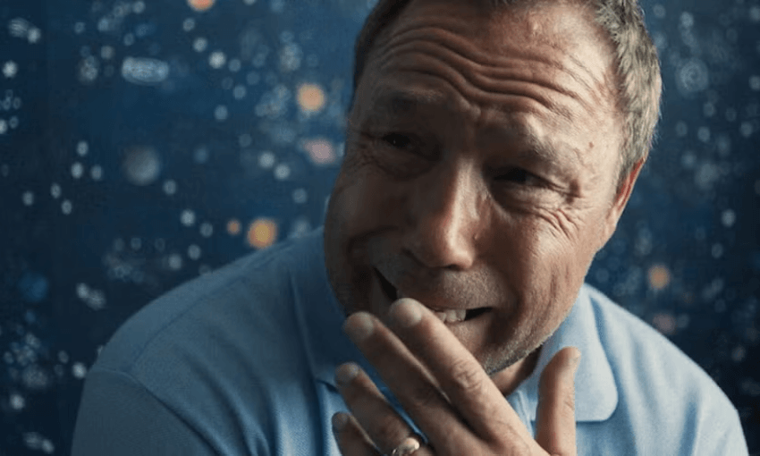 A man in a blue shirt (Stephen Graham as Ediie in Netflix's Adolescence) appears emotionally distraught, covering his mouth with his hand. The background is dark blue with a wallpaper pattern behind him resembling stars and galaxies.