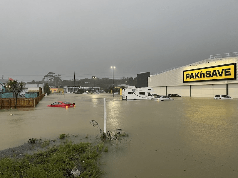 Flooded parking lot with submerged cars near a PAK'nSAVE store. The sky is overcast, and water levels are high, covering most of the vehicles, including a red car and a white van. Buildings and a wooden fence are partially visible in the background.