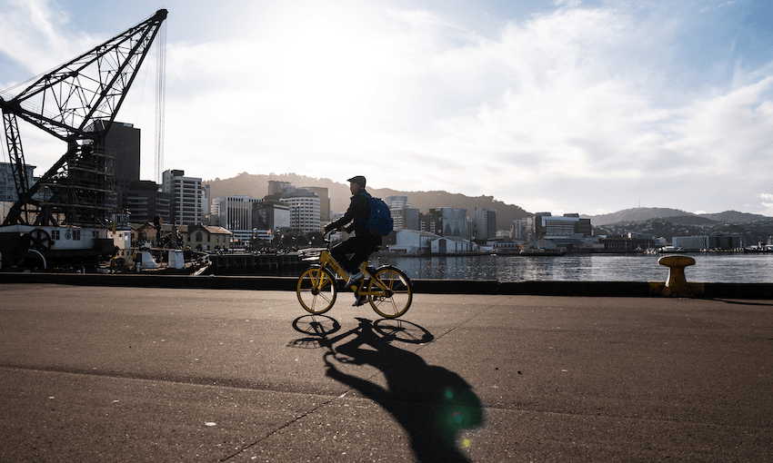 Person riding a yellow bike on a waterfront promenade with city buildings and hills in the background. The sun is shining brightly, casting a long shadow. An industrial crane stands to the left. The scene is set against a partly cloudy sky.