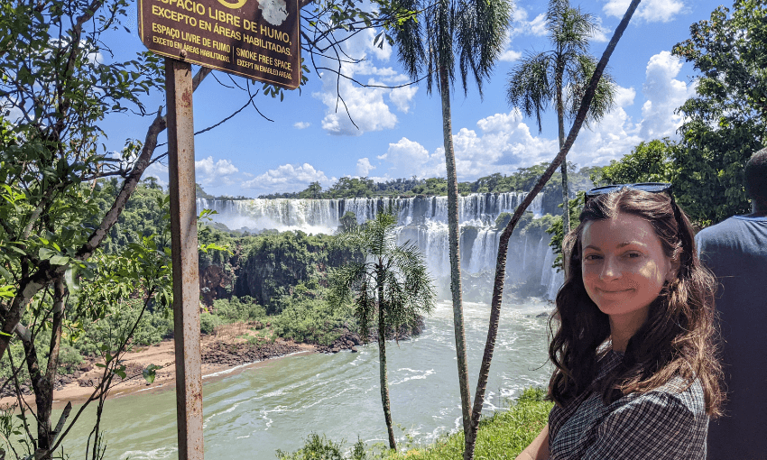 A woman stands in front of the Iguazu Falls, surrounded by lush greenery under a bright blue sky. A sign in Spanish nearby indicates a smoke-free area.