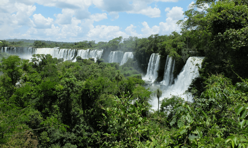 A lush jungle landscape with multiple cascading waterfalls, a section of Iguazu Falls, is surrounded by dense greenery under a partly cloudy sky.