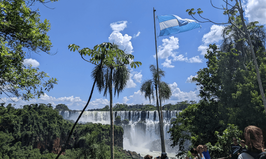 A scenic view of Iguazú Falls cascading behind lush green foliage under a bright blue sky. An Argentine flag waves in the foreground, with tourists observing the scene. Puffy clouds hang overhead, adding to the picturesque setting.