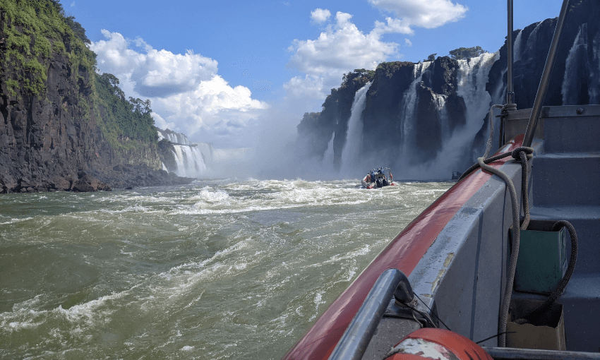 A boat navigates the rapids near the base of a large waterfall, with cascading water surrounded by lush green cliffs under a cloudy blue sky. The scene is viewed from another boat's perspective.