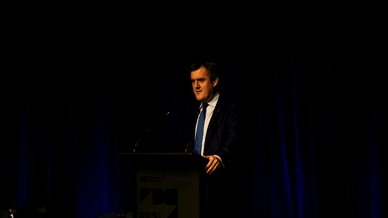 A man in a suit speaks at a podium on a dimly lit stage, with a dark blue curtain backdrop.