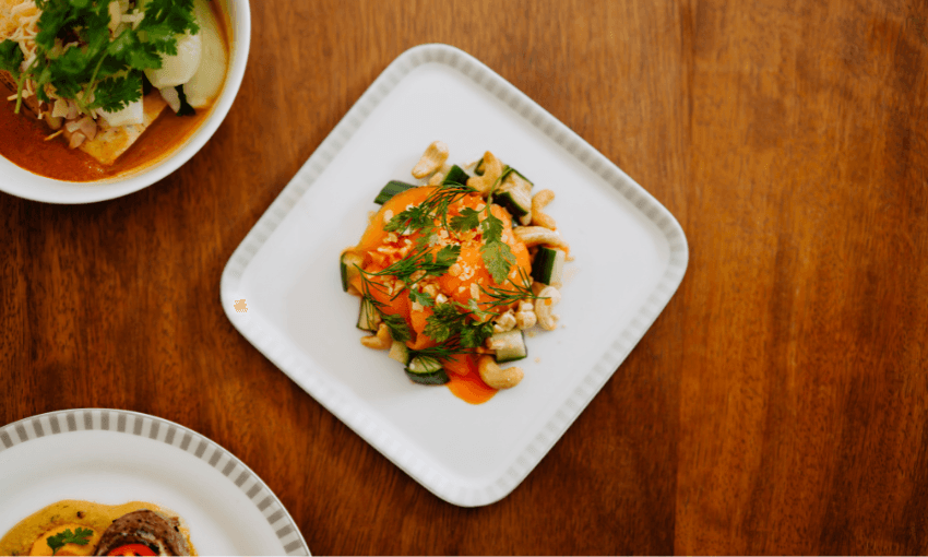 A square white plate with a dish featuring vegetables, greens, sauce, and nuts on a wooden table. Partially visible adjacent dishes suggest a meal setting.
