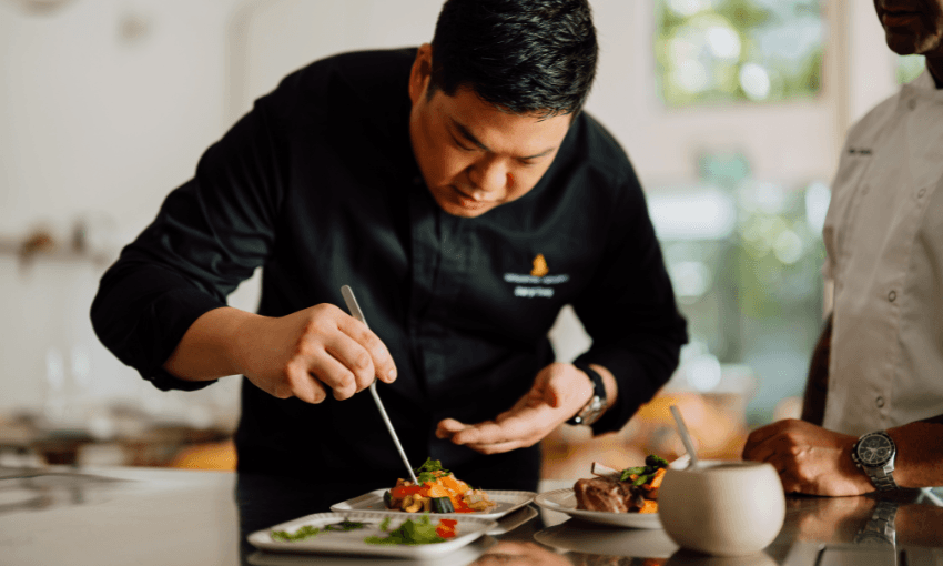 A chef in a black uniform is carefully plating a dish in a bright kitchen. Another person in a white chef jacket observes. Fresh ingredients and small plates are visible on the counter.