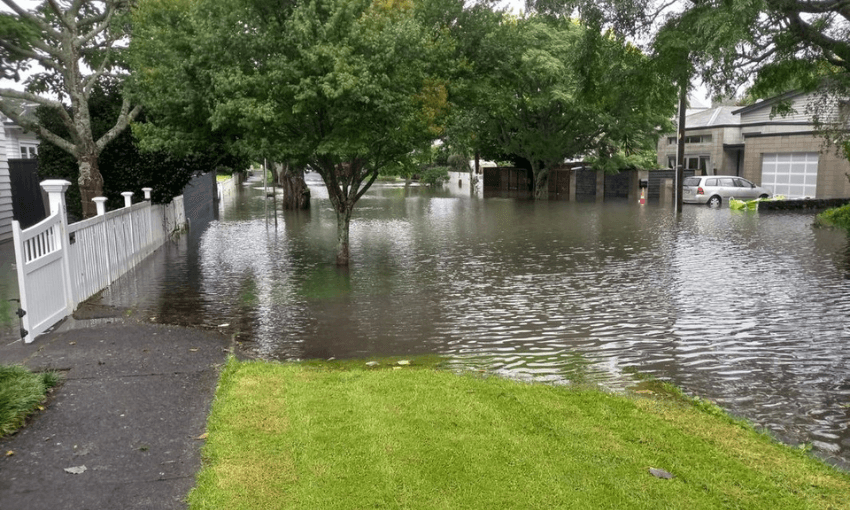 A flooded suburban street with water covering the road, reaching up to the lawns of houses. Trees are partially submerged, and a car is visible near a house. Overcast sky suggests recent heavy rain.