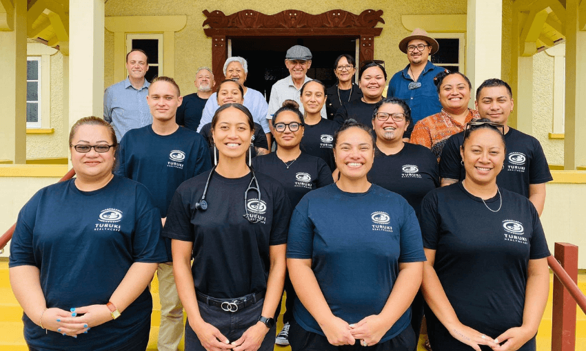 A group of people and doctors from Turuki Healthcare is posing on the steps of a building. The front row consists of eight individuals wearing black shirts with a logo. Behind them, others are dressed casually. The building features a decorative entrance with yellow and red details.