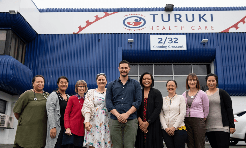A group of people standing in front of Turuki Healthcare and smiling in front of a building with "Turuki Health Care" signage. The building is blue and white, located at 2/32 Canning Crescent. The group appears cheerful and casually dressed.