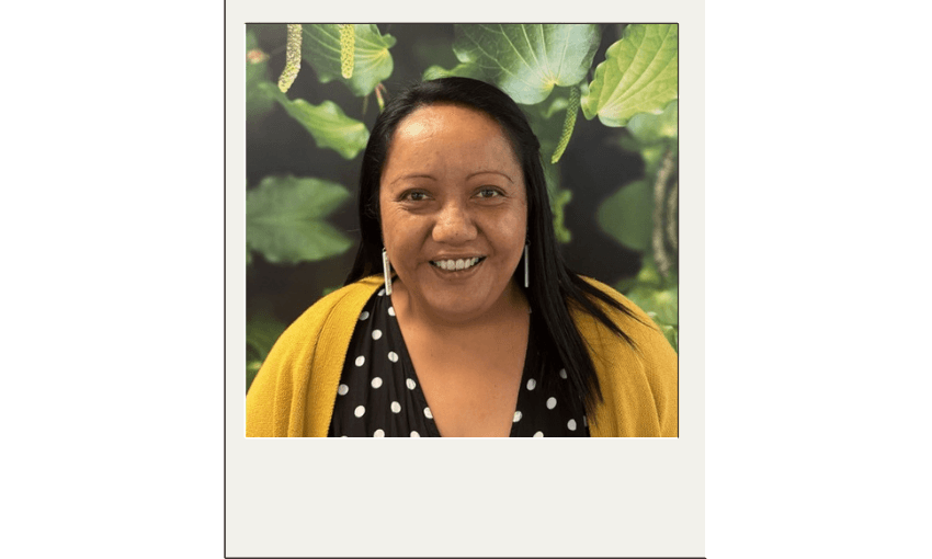 Priscilla Laita, a woman with long black hair smiles at the camera. She is wearing a black top with white polka dots and a mustard yellow cardigan. The background features large green kawakawa leaves and berries.