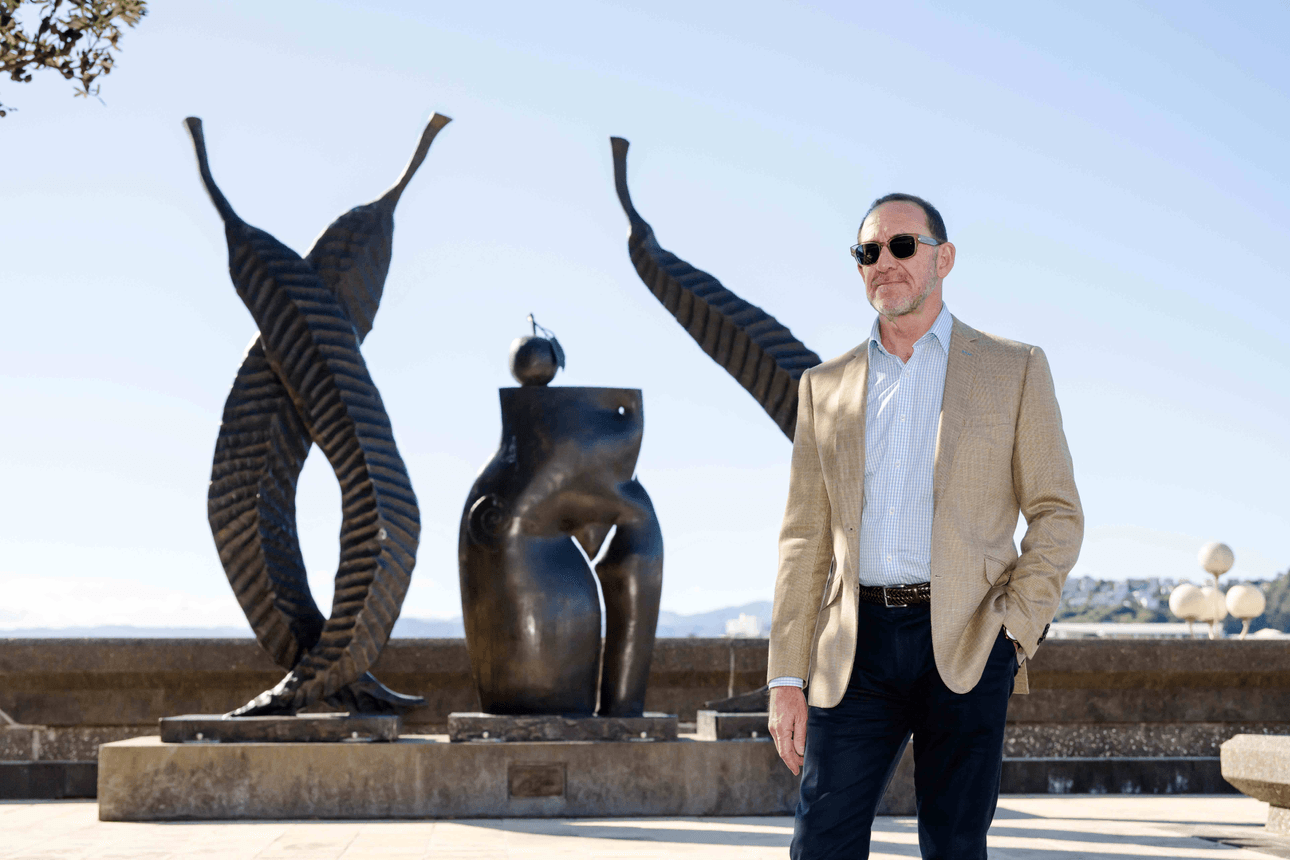 A man in sunglasses and a beige blazer stands in front of abstract bronze sculptures on an outdoor plaza, with a clear sky and cityscape visible in the background.