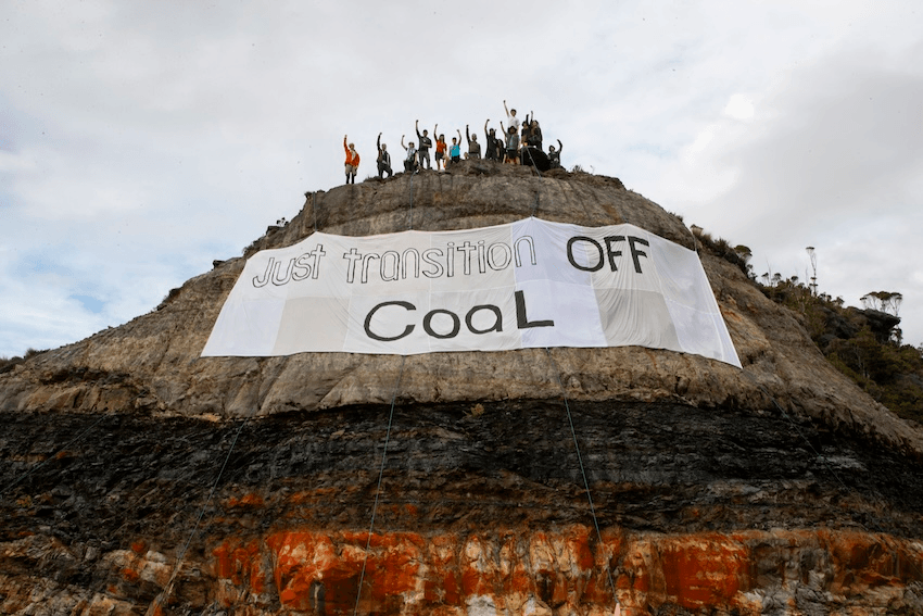 a group of people on top of a hill with a large banner hanging down over the exposed earth saying 'just transition off coal" 