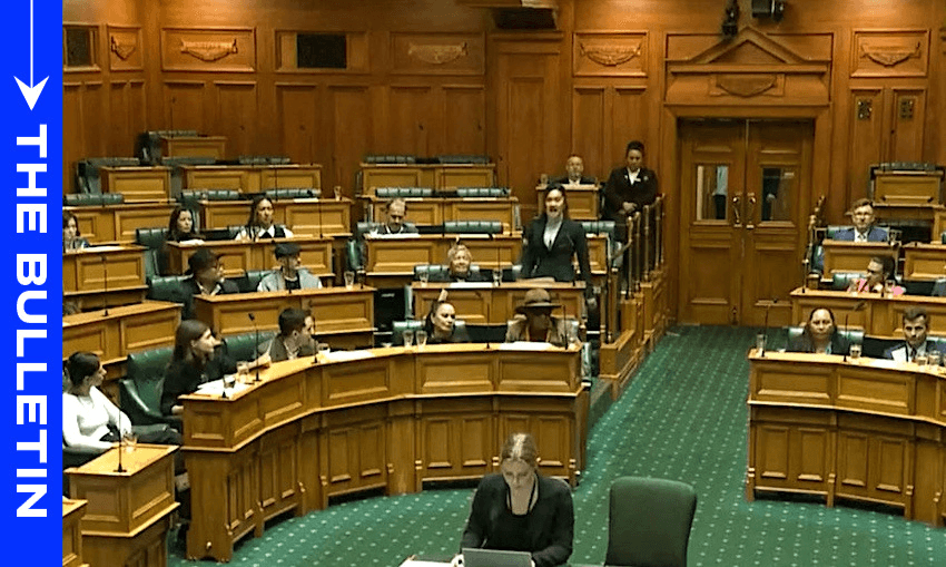 People are seated in a parliamentary chamber with wooden interiors and green seats. A person is standing and speaking, while others listen. A large screen displays the text "The Bulletin" on the left side.
