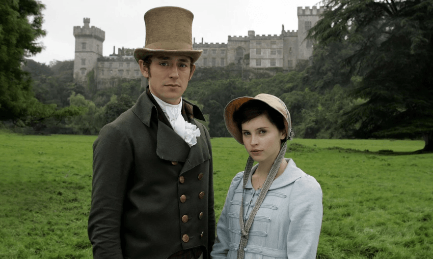 Felicity Jones as Catherine Morland and JJ Feild as Henry Tilney in the 2007 film of Northanger Abbey. They are standing in costume on a grassy field in front of a huge Abbey.