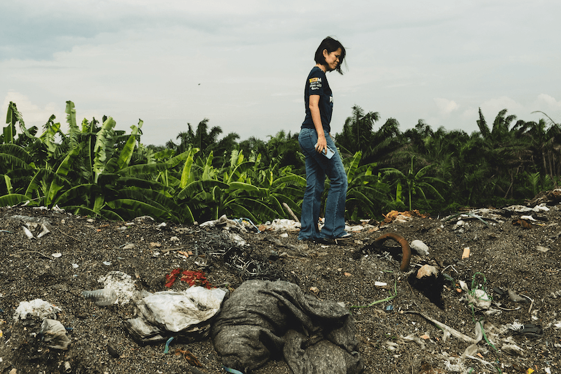 A person walks on a mound of dirt and scattered trash, holding a white object. They are surrounded by green banana trees under a cloudy sky. The scene suggests a focus on environmental pollution or waste management.