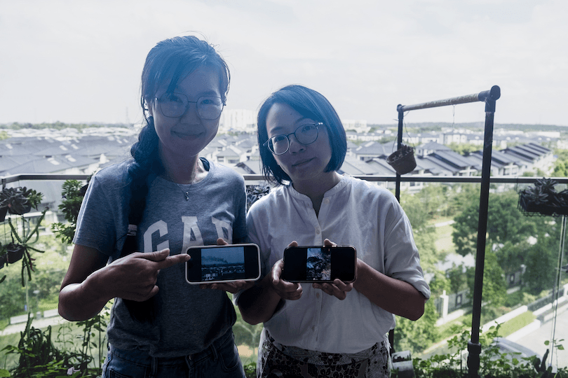 Two women stand on a balcony, each holding a smartphone. The woman on the left points to her phone, and both phones display images. Behind them is a view of rooftops and greenery under a cloudy sky.
