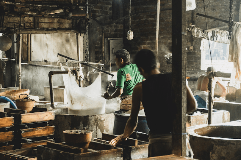 Workers in a rustic workshop setting, engaged in processing materials using traditional methods. The room is filled with wooden equipment and steam, creating a warm, industrious atmosphere.