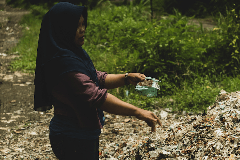 A person wearing a dark headscarf stands by a large pile of trash, holding a mask in one hand, in an outdoor setting surrounded by greenery. The scene conveys an environmental concern.