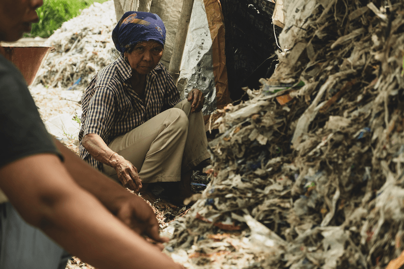 An elderly person wearing a headscarf sits near a large pile of mixed waste, sorting through it. Another person is visible in the foreground, partially seen, engaging in a similar activity. The scene suggests recycling or waste sorting.